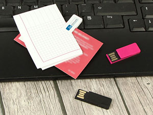 USB-Sticks in pink und schwarz liegen auf einer Tastatur, umgeben von Notizkarten und einem farbigen Blatt.