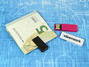 USB-Sticks in verschiedenen Farben liegen neben einem 5-Euro-Schein auf einem blauen Hintergrund mit technischen Zeichnungen.