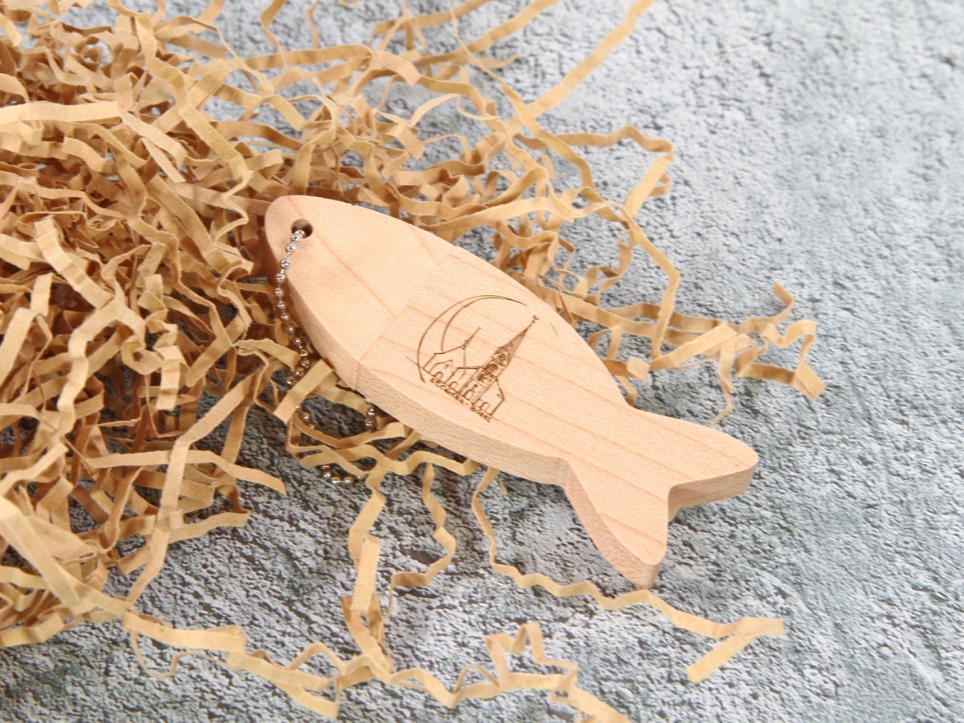 USB-Stick in Fischform aus Holz mit Platz für individuelles Logo, präsentiert auf dekorativem Füllmaterial.